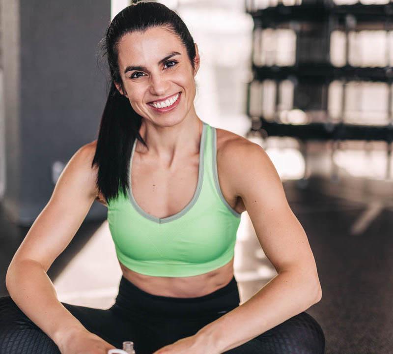 Beautiful young female in sportswear smiling and looking at camera while sitting on floor on blurred background of gym