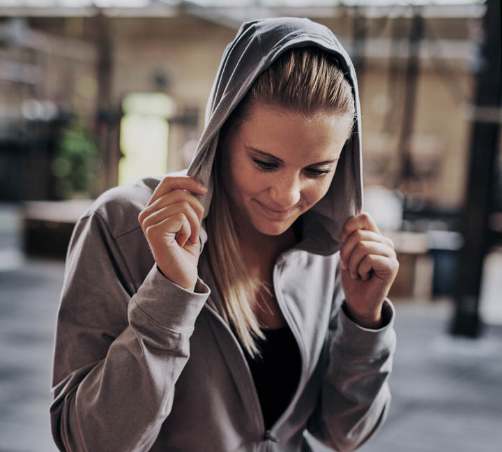 Sporty young blonde woman smiling and putting on the hood of her sweatshirt while sitting on a box after a workout at the gym